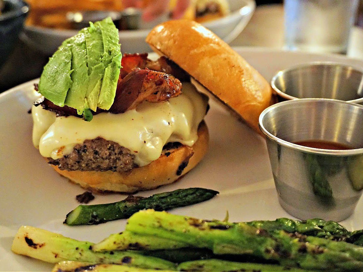 Where To Dine In the Shoals Cheeseburger topped with avocado and bacon and served with a side of asparagus.