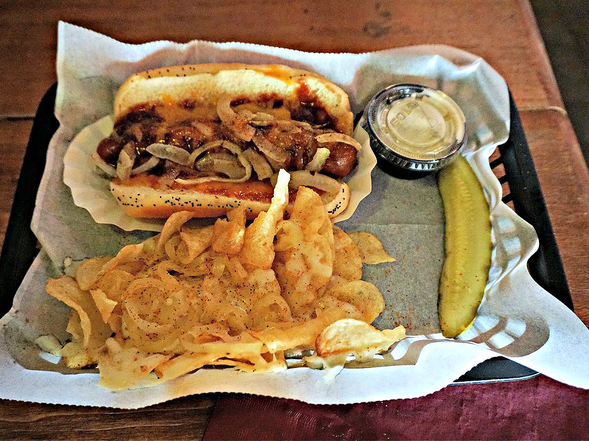 Where To Dine In the Shoals Deluxe hot dog on a tray with a side of potato chips and a pickle spear.