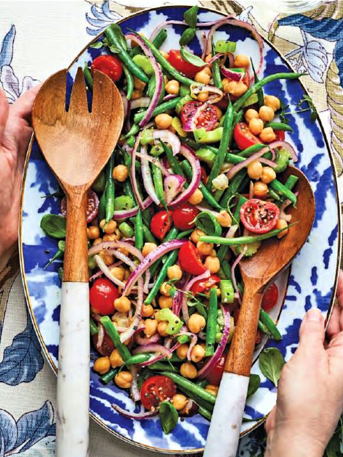 Woman's hands holding a blue and white platter of vegetable salad and wooden utensils.