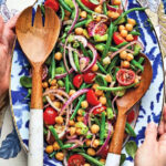 Woman's hands holding a blue and white platter of vegetable salad and wooden utensils.