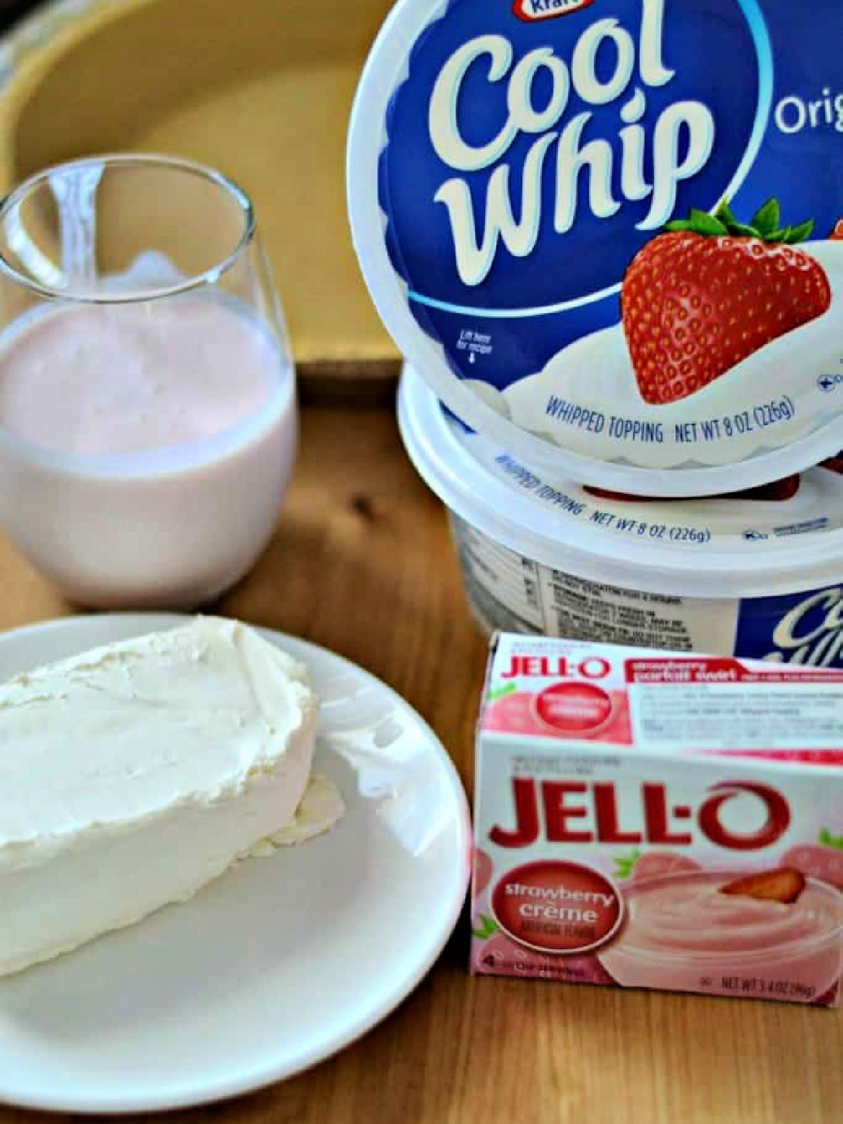Block of cream cheese, strawberry kefir, strawberry pudding mix, and two containers of Cool Whip on a cutting board.