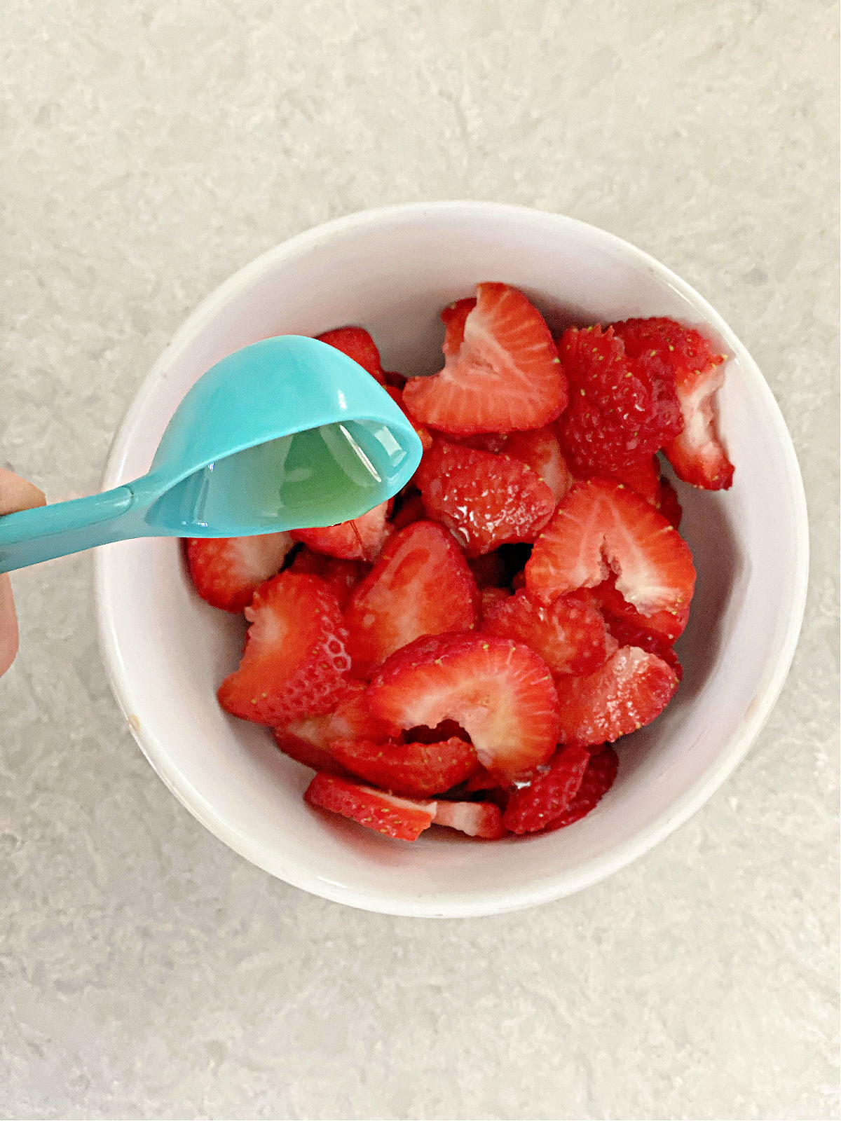 Blue tablespoon with maple syrup being poured onto bowl of strawberries.