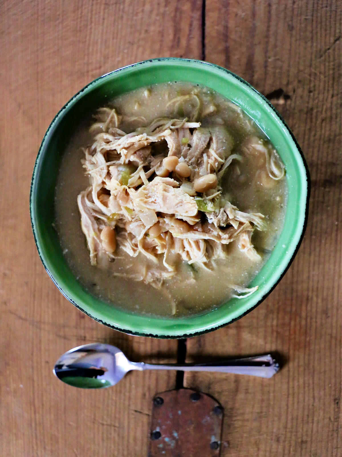 White chicken chili in a green bowl with a spoon on a wooden board.
