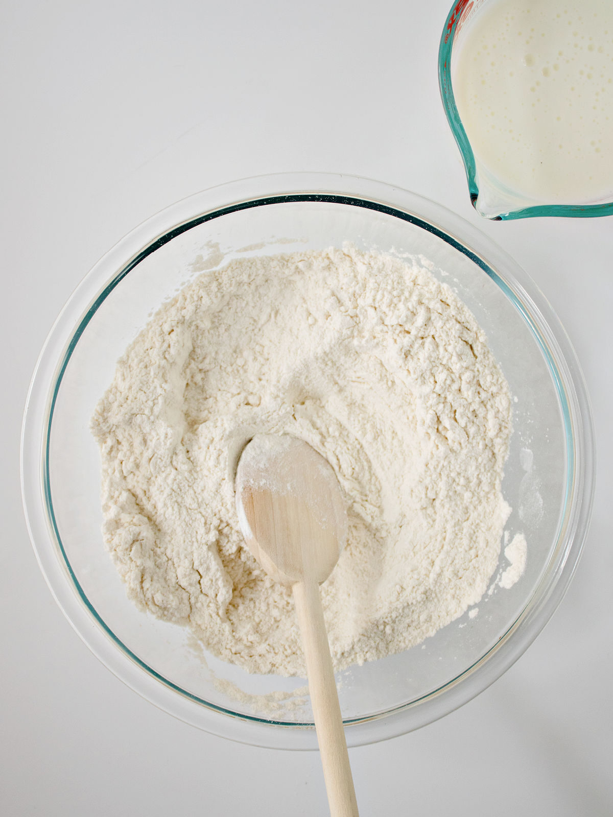 Glass bowl with flour and wooden spoon by a measuring cup with buttermilk.