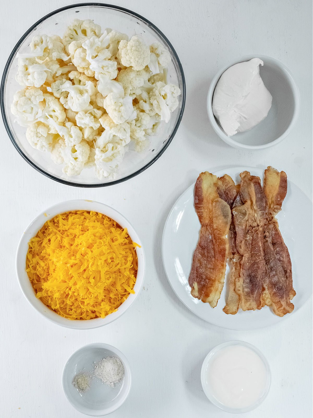 Bowl of cauliflower, bowl of Cheddar cheese, ramekin with cream cheese, plate of bacon, and ramekins of seasonings.