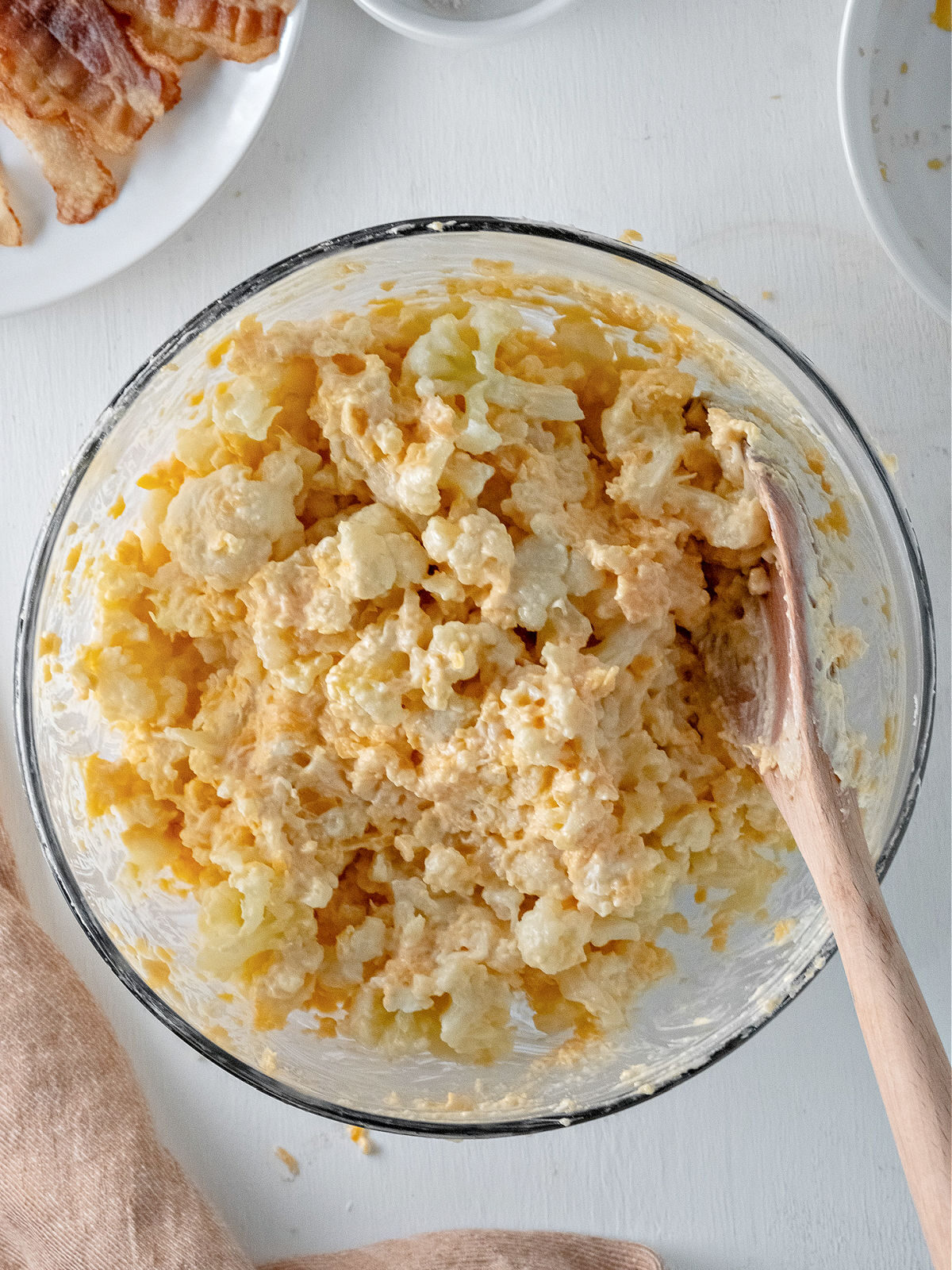 Glass bowl filled with cauliflower and cheese being stirred with a wooden spoon.