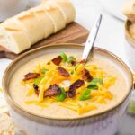 Bowl of potato soup topped with bacon, cheese, and chives with loaf of bread.