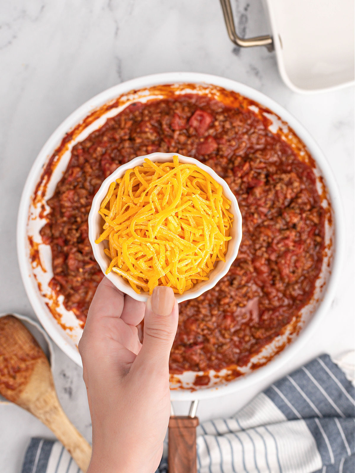 Woman's hand holding bowl of Cheddar cheese over cooked ground beef.