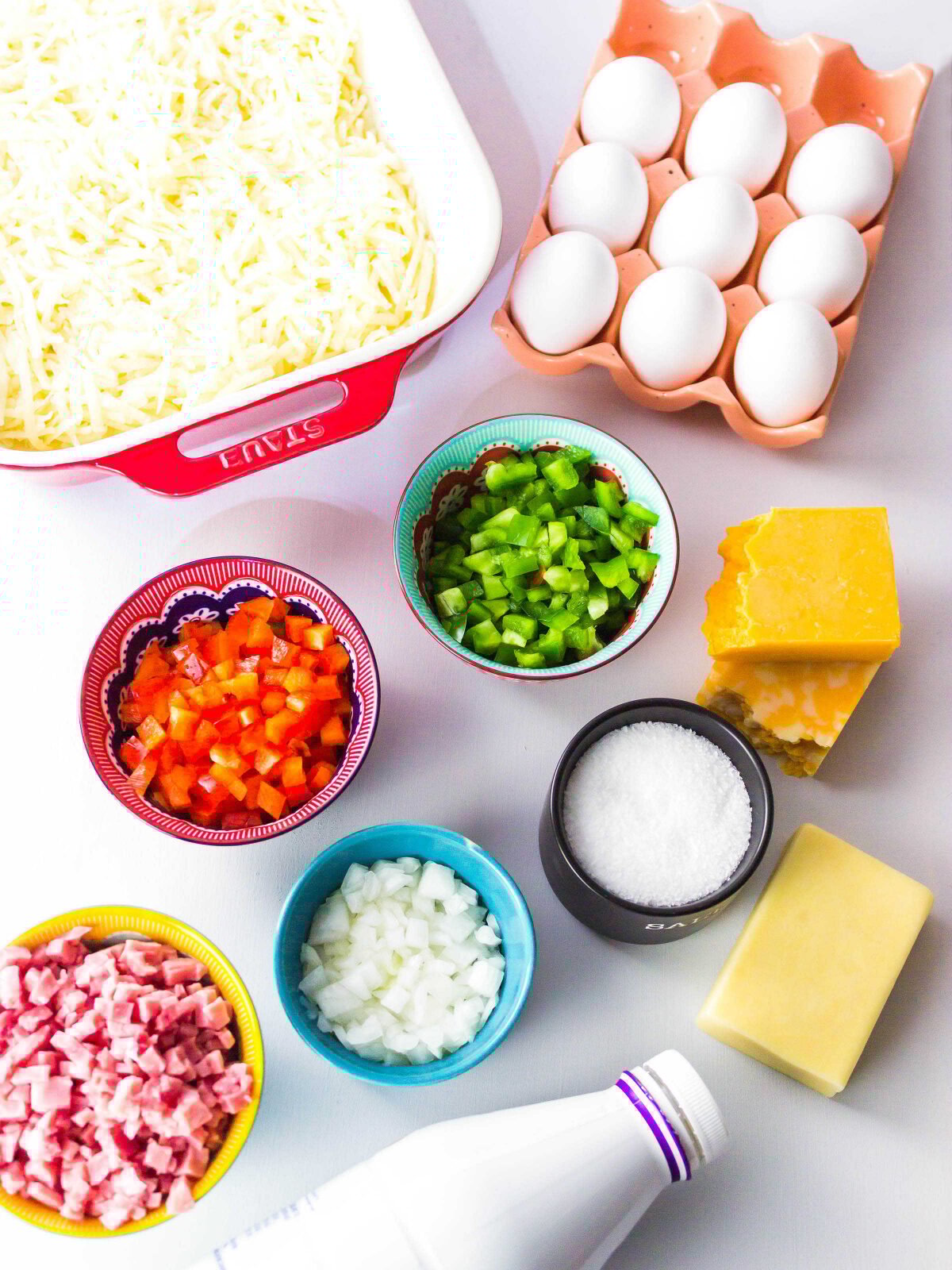 Hash brown potatoes in a baking dish, eggs, diced peppers, diced onion and ham in bowls.