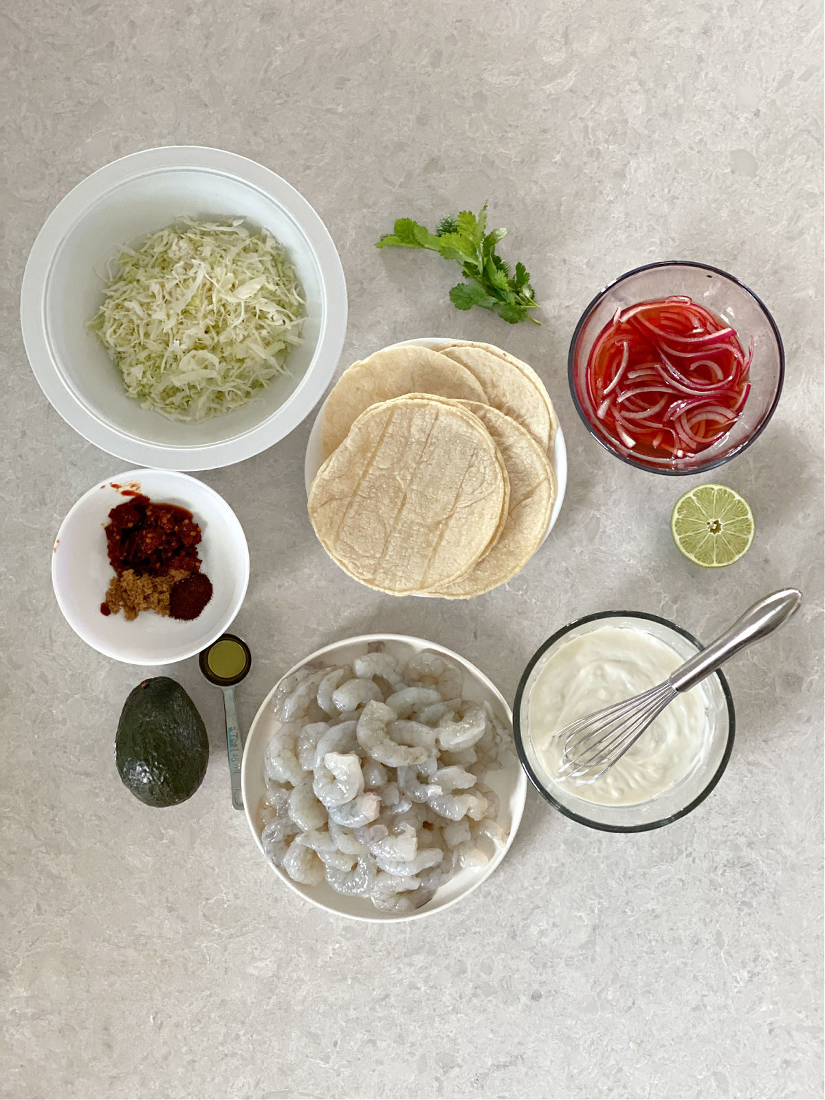 Shredded cabbage, seasoning, raw shrimp, pickled onions, avocado, and yogurt dressing on a counter.