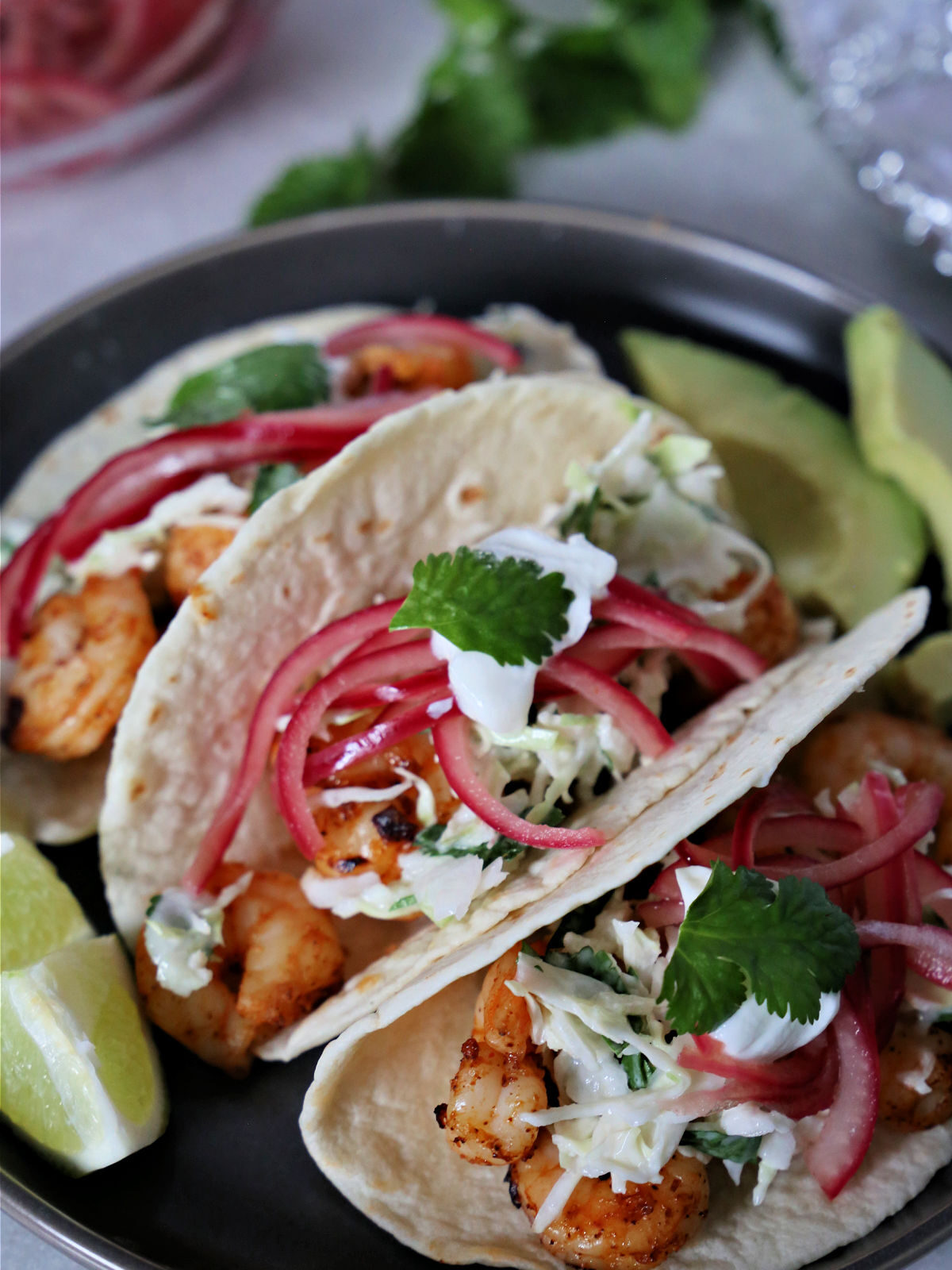 Three air fried shrimp tacos on a plate with avocado slices and lime wedges.