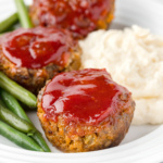 Three meatloaf muffins on a plate with green beans and mashed potatoes.