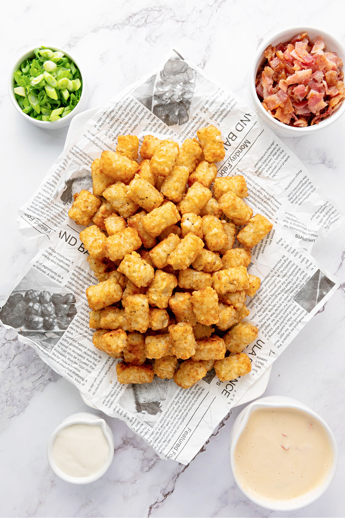 Baked tater tots surrounded by bowls with bacon, green onions, Ranch, and queso cheese.