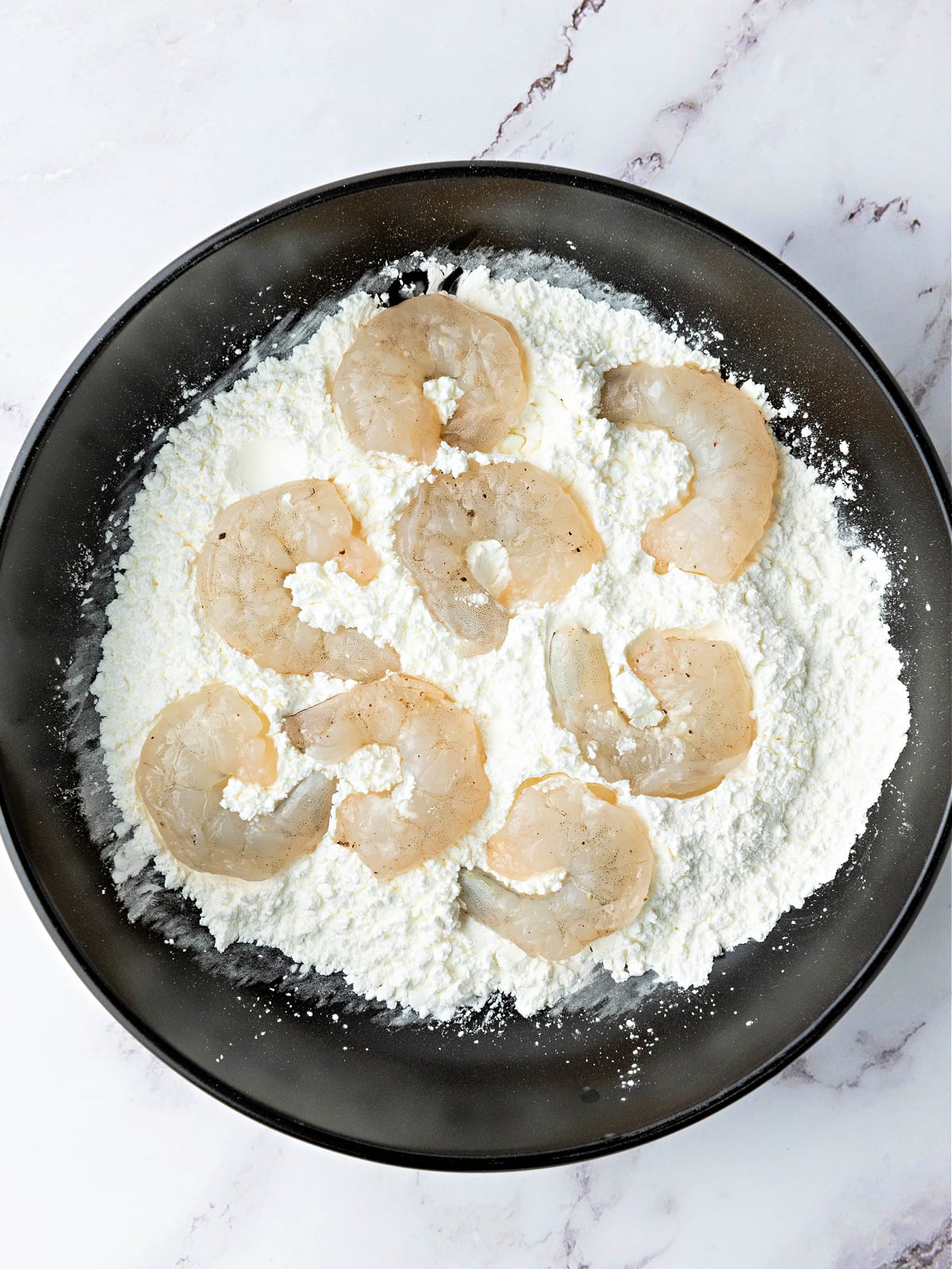 Shrimp on top of a flour and cornstarch mixture on a black plate.