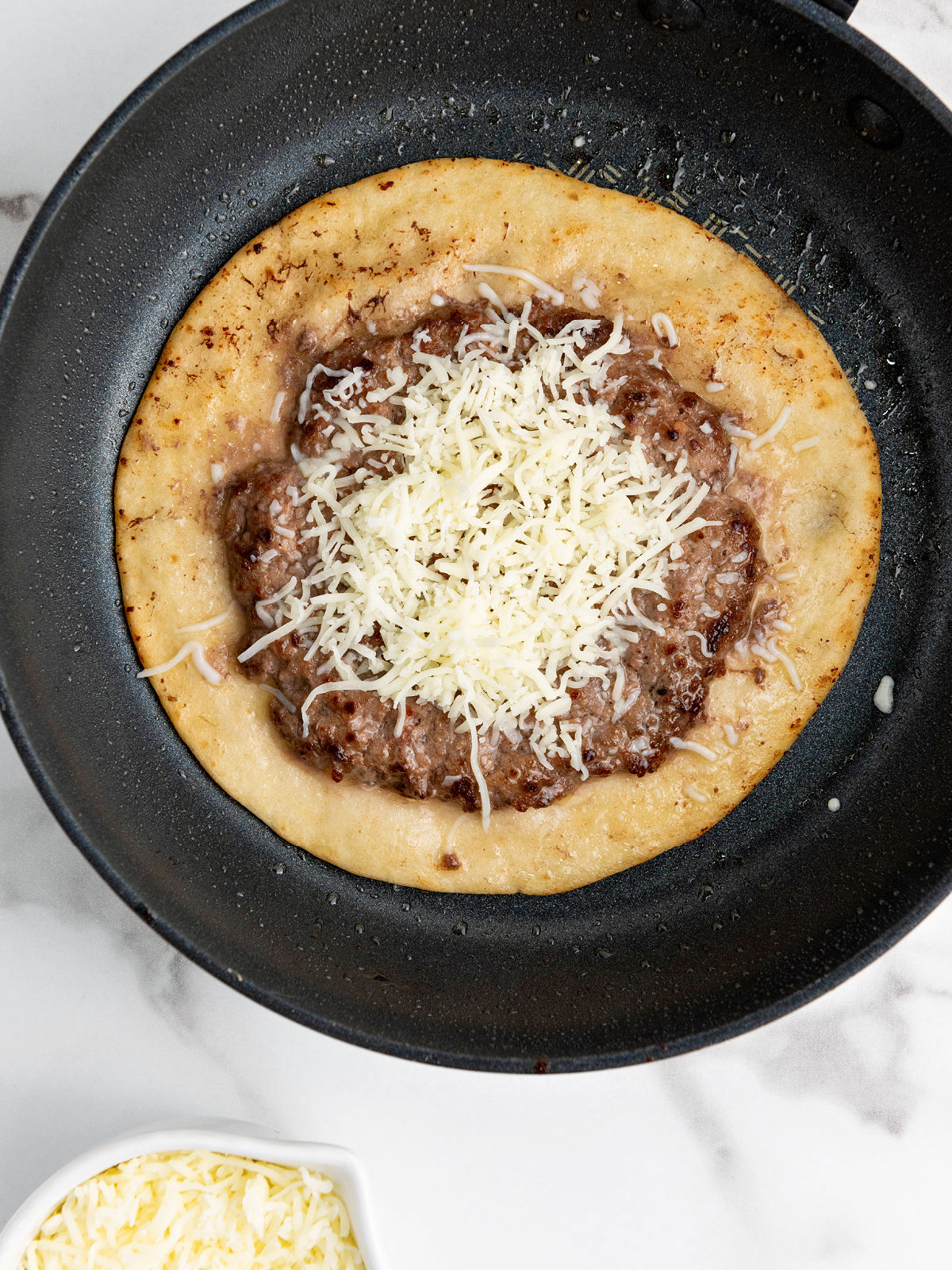 A tortilla in a skillet topped with cooked ground beef and shredded cheese.