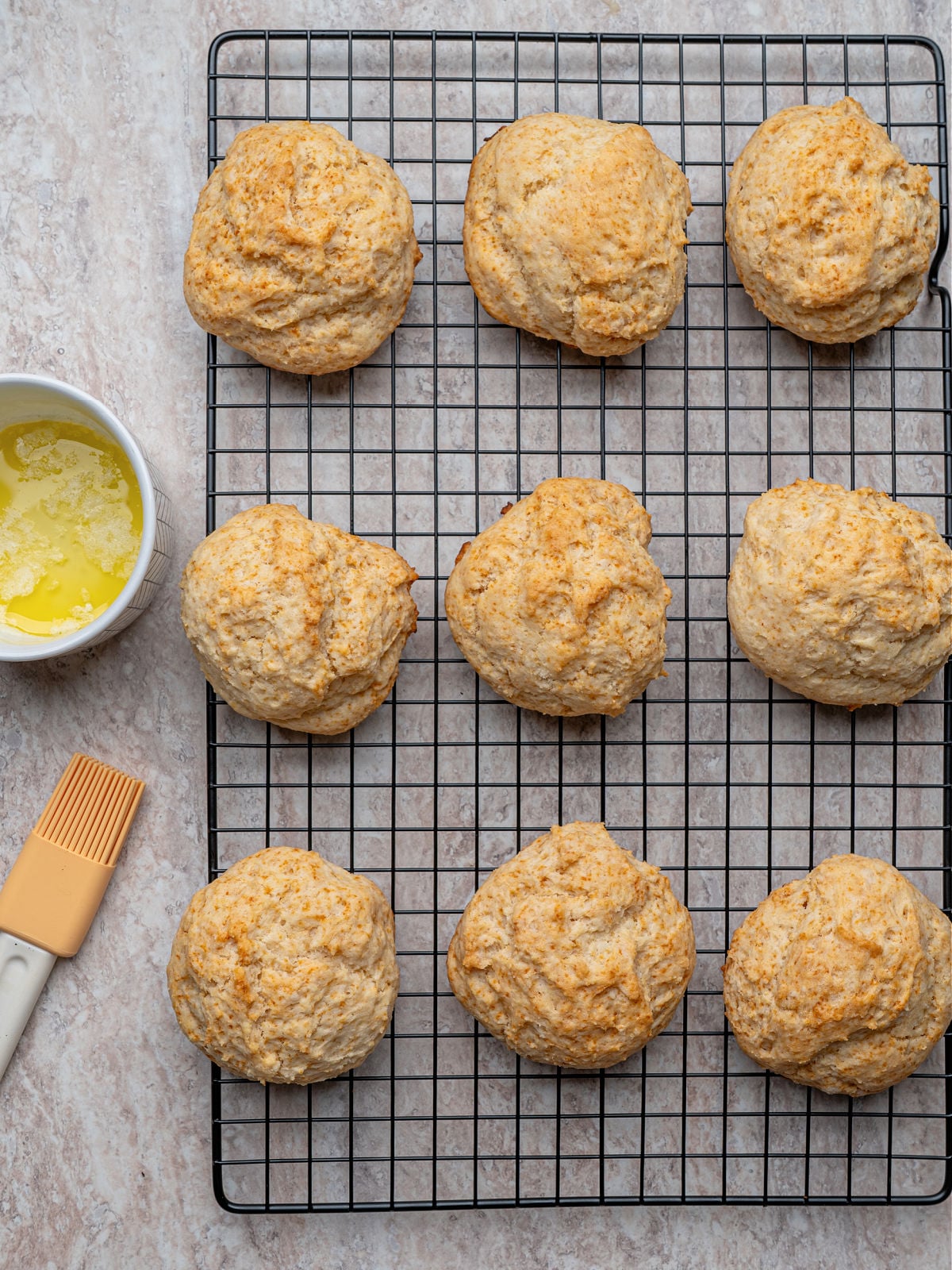 Golden-brown buttermilk drop biscuits cooling on a wire rack beside a small bowl of melted butter.