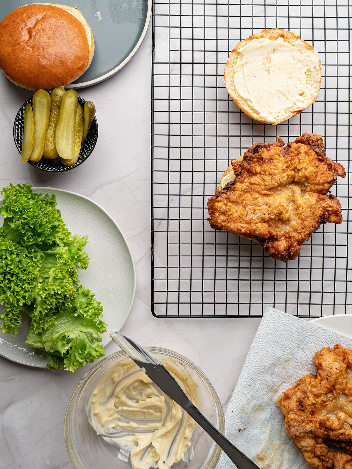 Fried chicken thigh on top of a brioche bun with aioli on one slice, a plate of curly lettuce, and a small bowl of pickles.