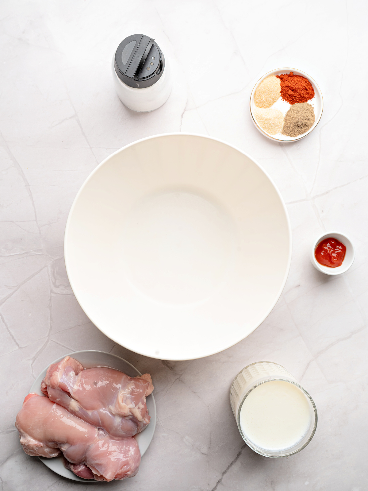 Chicken thighs, buttermilk, spices, and a large white bowl to be used to make fried chicken sandwiches.