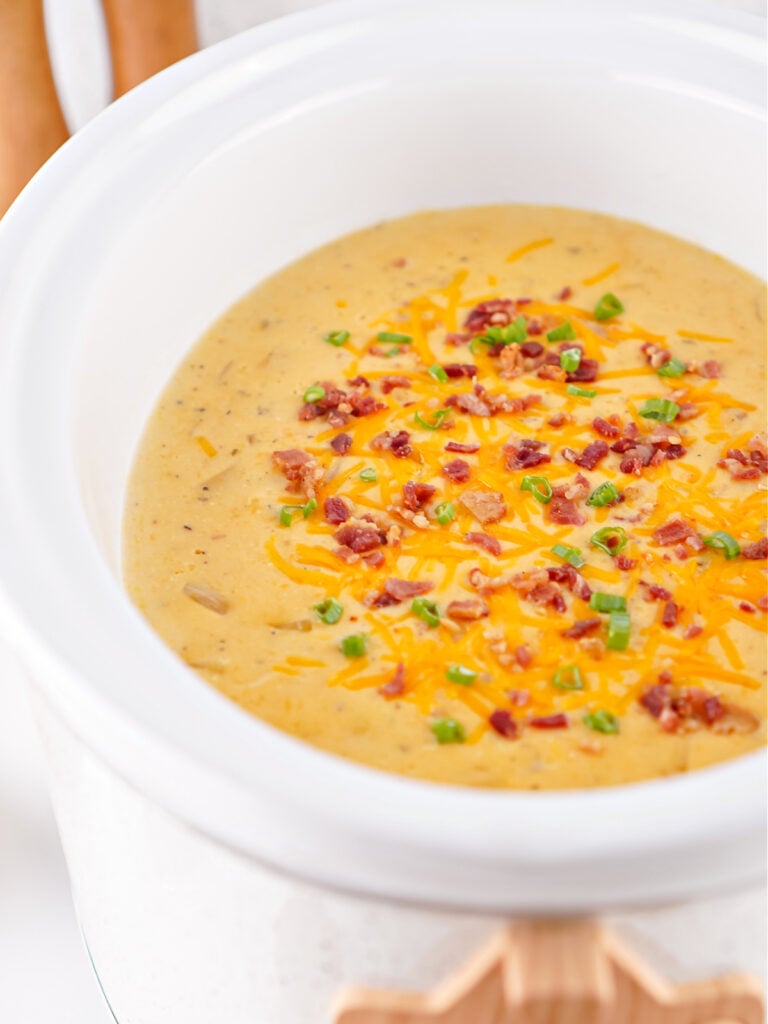 Crockpot filled with creamy potato soup, garnished with shredded cheese and crispy bacon.