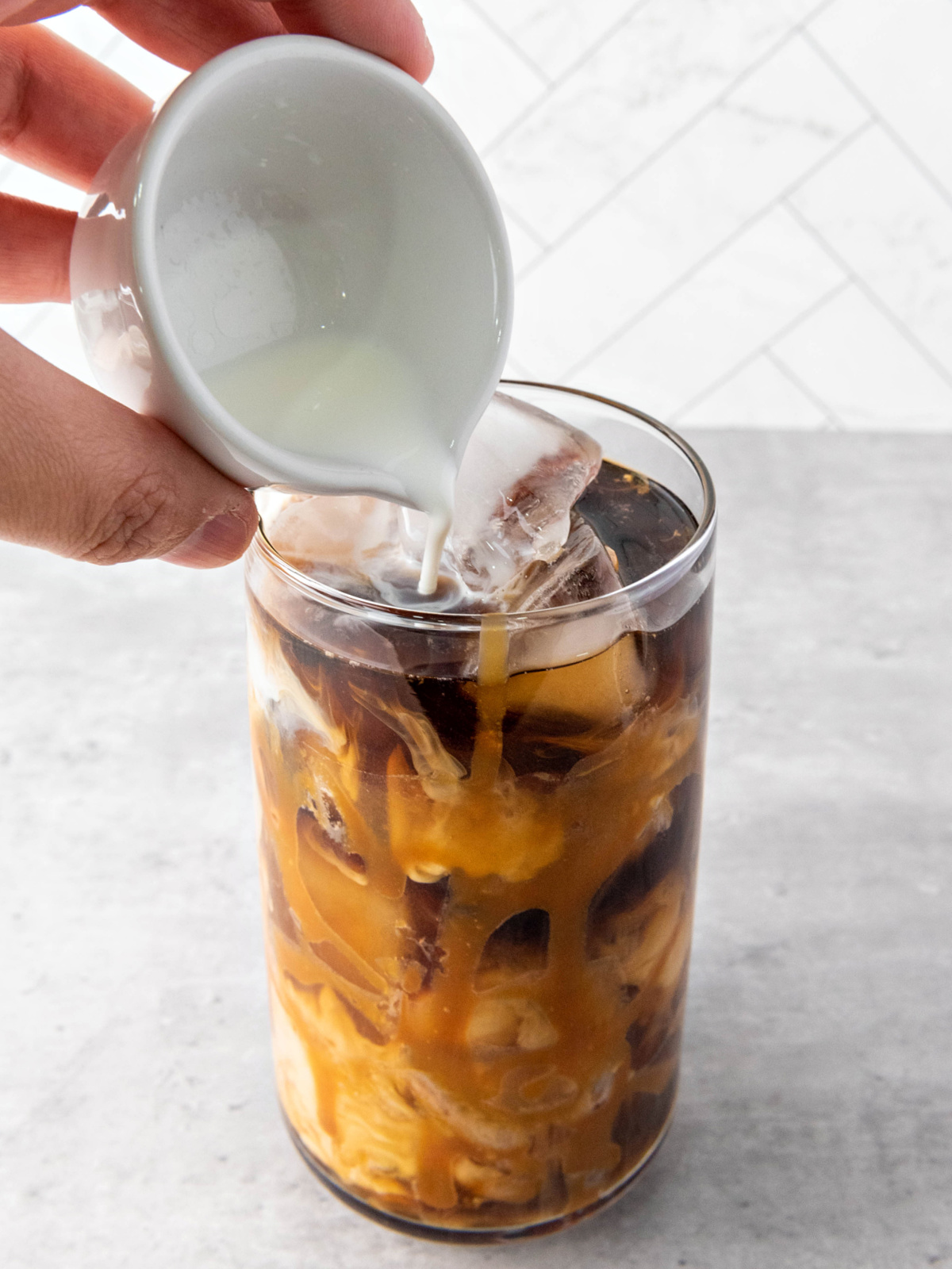 Half-and-half being poured into a glass of iced coffee, creating creamy caramel swirls.