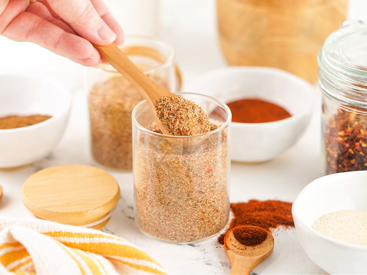 Hand holding a wooden spoon scooping homemade jerk seasoning from a jar.