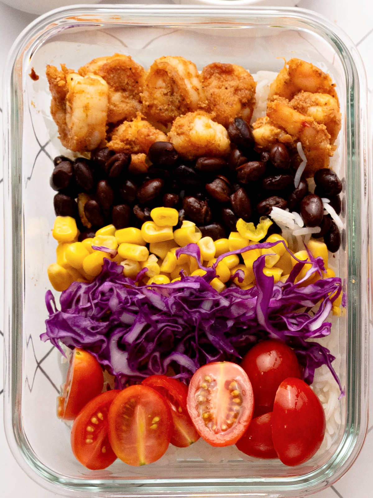 Seasoned shrimp, black beans, corn, cabbage, and tomatoes on a bed of rice in a glass bowl.