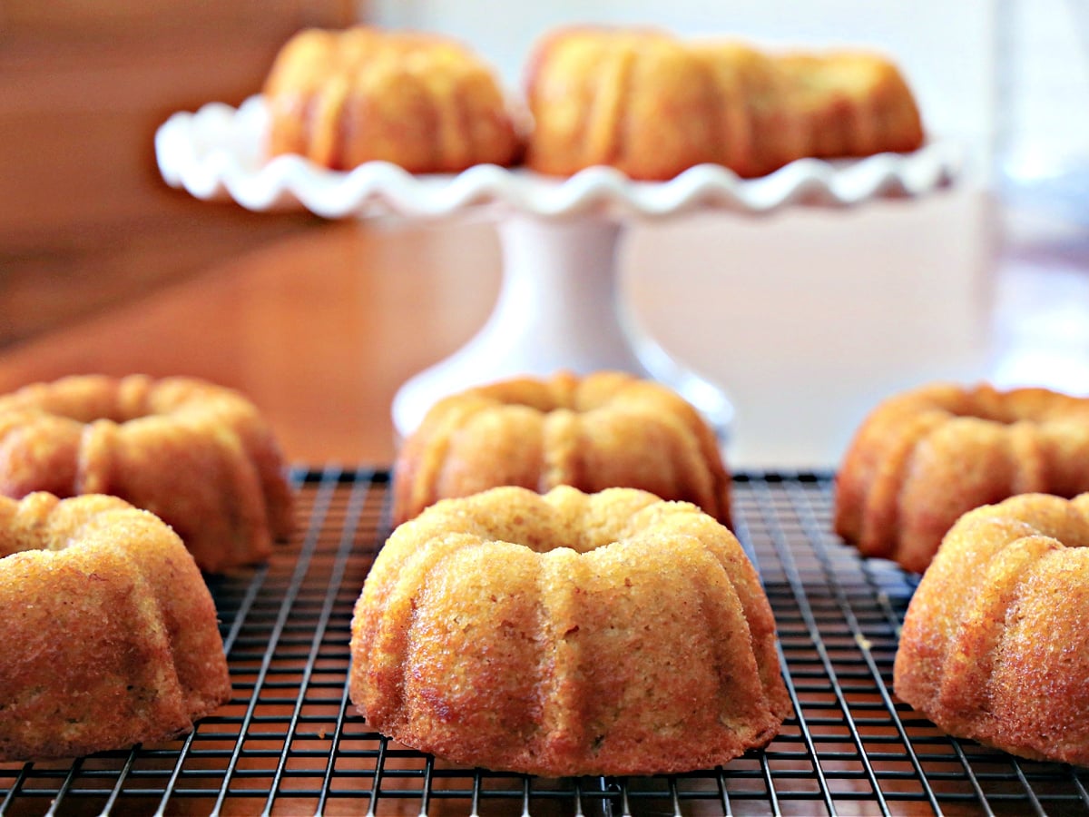 Six mini banana bundt cakes cooling on a wire rack with additional cakes displayed on a white pedestal stand in the background.