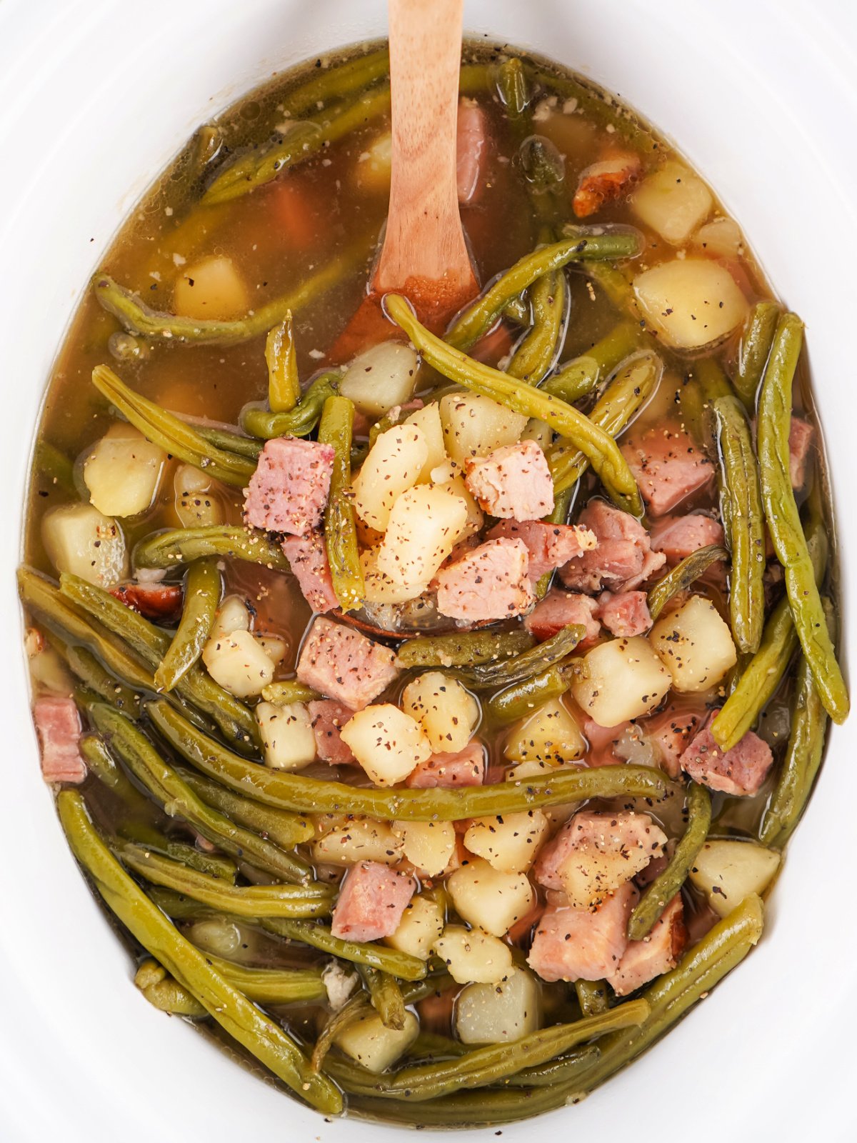 A spoon lifting a hearty serving of ham, green beans, and potatoes from a Crockpot, showing tender vegetables and savory chunks of ham.