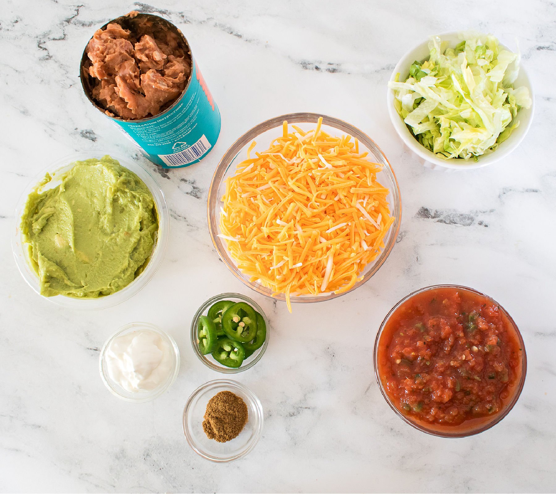 Ingredients for layered bean dip displayed on a marble countertop, including refried beans, guacamole, shredded lettuce, shredded cheese, salsa, sour cream, cumin, and sliced jalapeños.