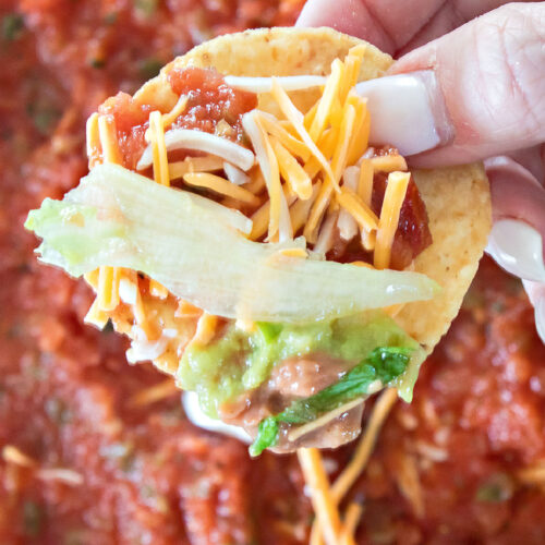 Hand holding a tortilla chip loaded with layered bean dip including beans, guacamole, lettuce, shredded cheese, salsa, and sour cream.