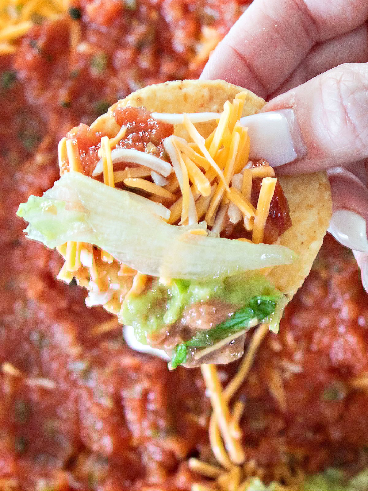 Hand holding a tortilla chip loaded with layered bean dip including beans, guacamole, lettuce, shredded cheese, salsa, and sour cream.