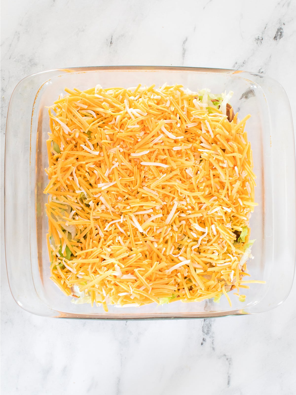 Glass baking dish filled with layered bean dip topped generously with shredded Mexican cheese on a marble surface.
