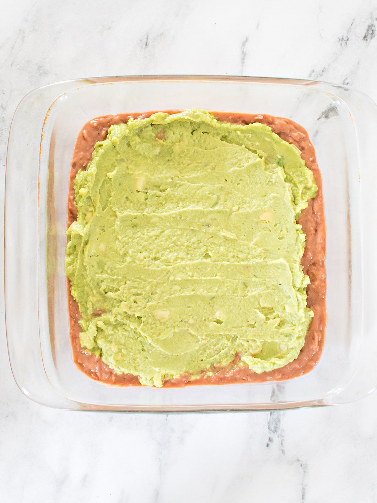 Clear baking dish with refried beans spread on the bottom and a thick layer of guacamole on top.