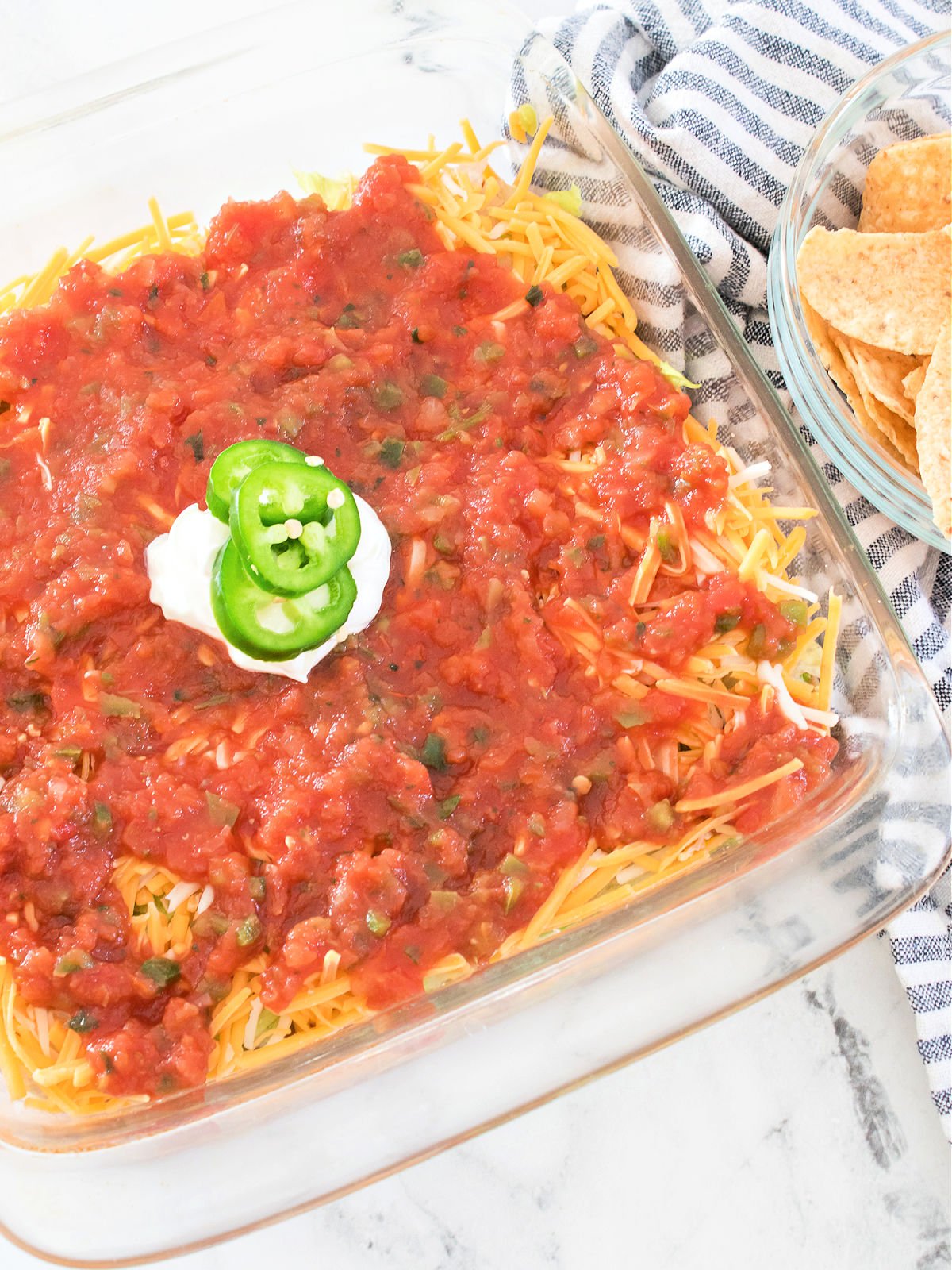 Layered bean dip in a glass dish topped with salsa, a dollop of sour cream, and sliced jalapeños, next to a bowl of tortilla chips.