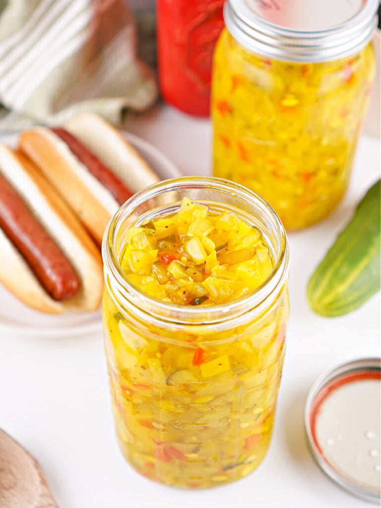 Two jars of homemade sweet pickle relish arranged on a white surface with fresh cucumber and two hotdogs nearby.
