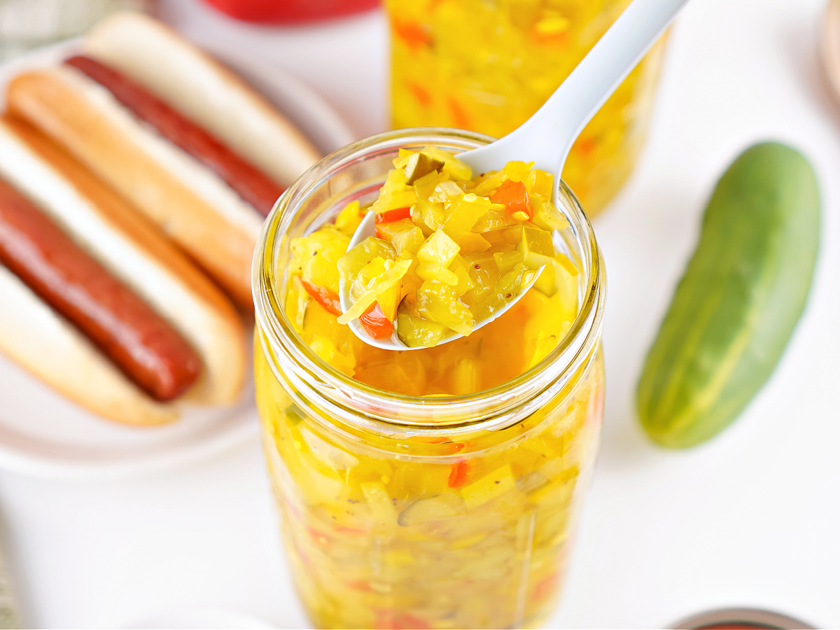 Close-up of sweet pickle relish on a spoon above an open jar, showing chopped cucumbers and peppers.