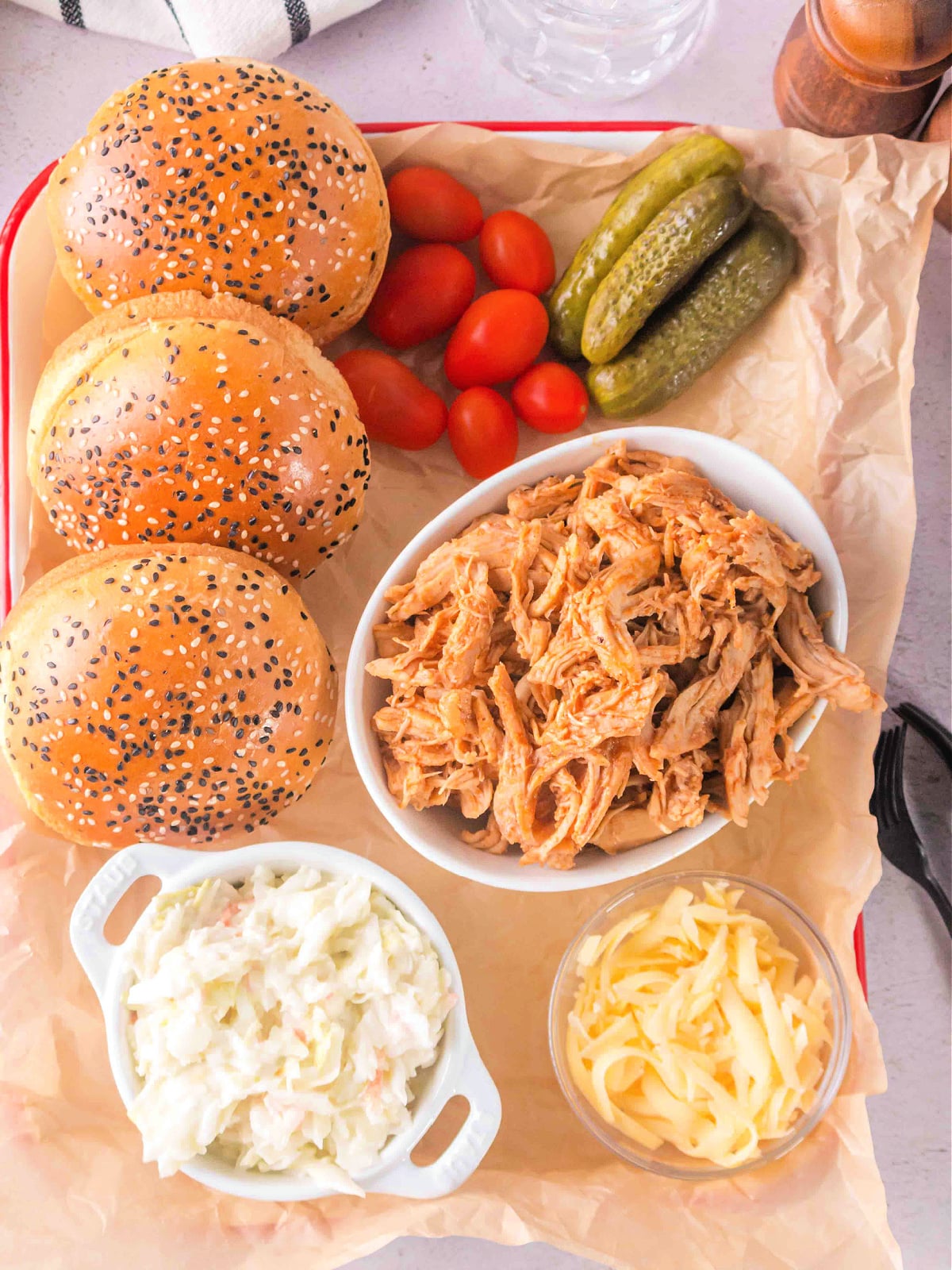 Overhead view of BBQ shredded chicken with sandwich buns, coleslaw, sliced pickles, cherry tomatoes, and shredded cheese on parchment paper.