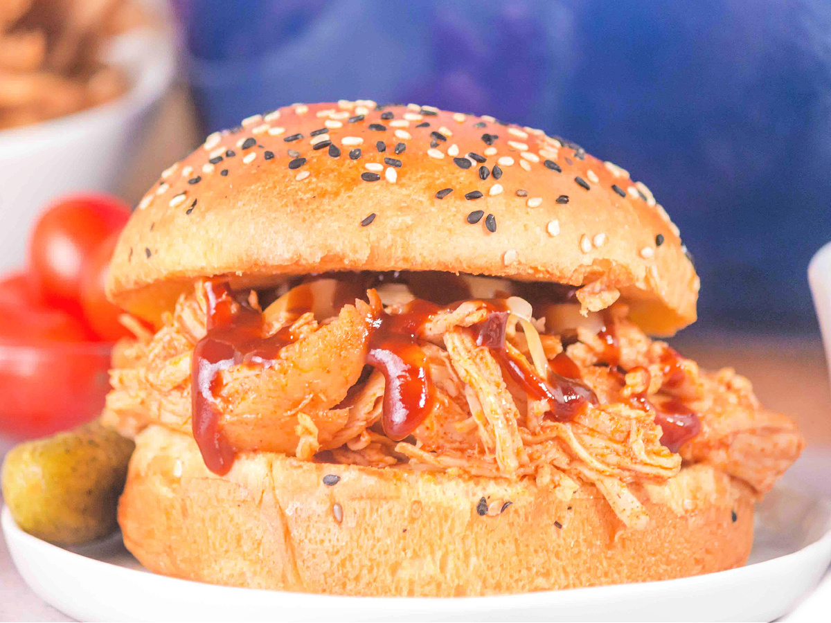 BBQ shredded chicken sandwich on a seeded bun topped with cheese and sauce, served with pickles.