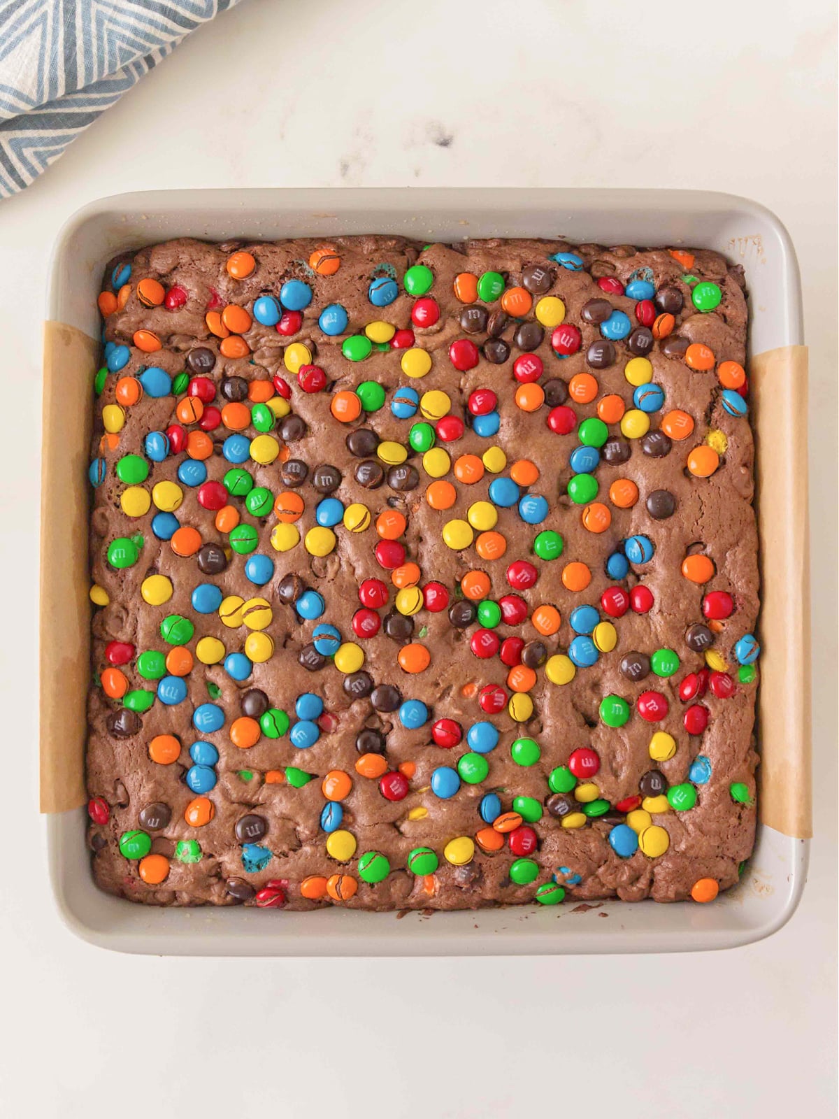 Overhead view of baked chocolate brownies topped with colorful M&M candies in a parchment-lined baking dish.