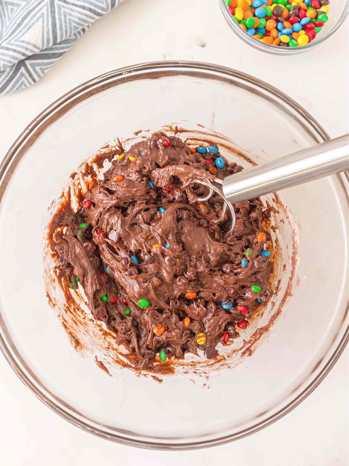 Thick chocolate brownie batter mixed with chocolate chips and M&M candies in a glass bowl.