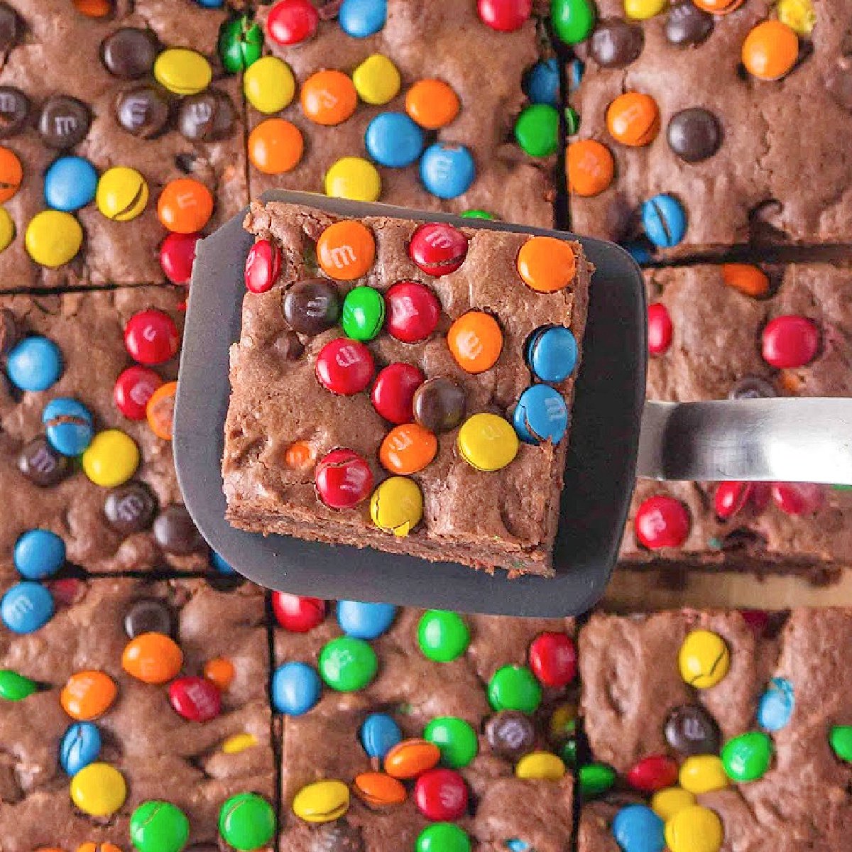 A square of M&M brownie lifted on a spatula showing a fudgy chocolate center.