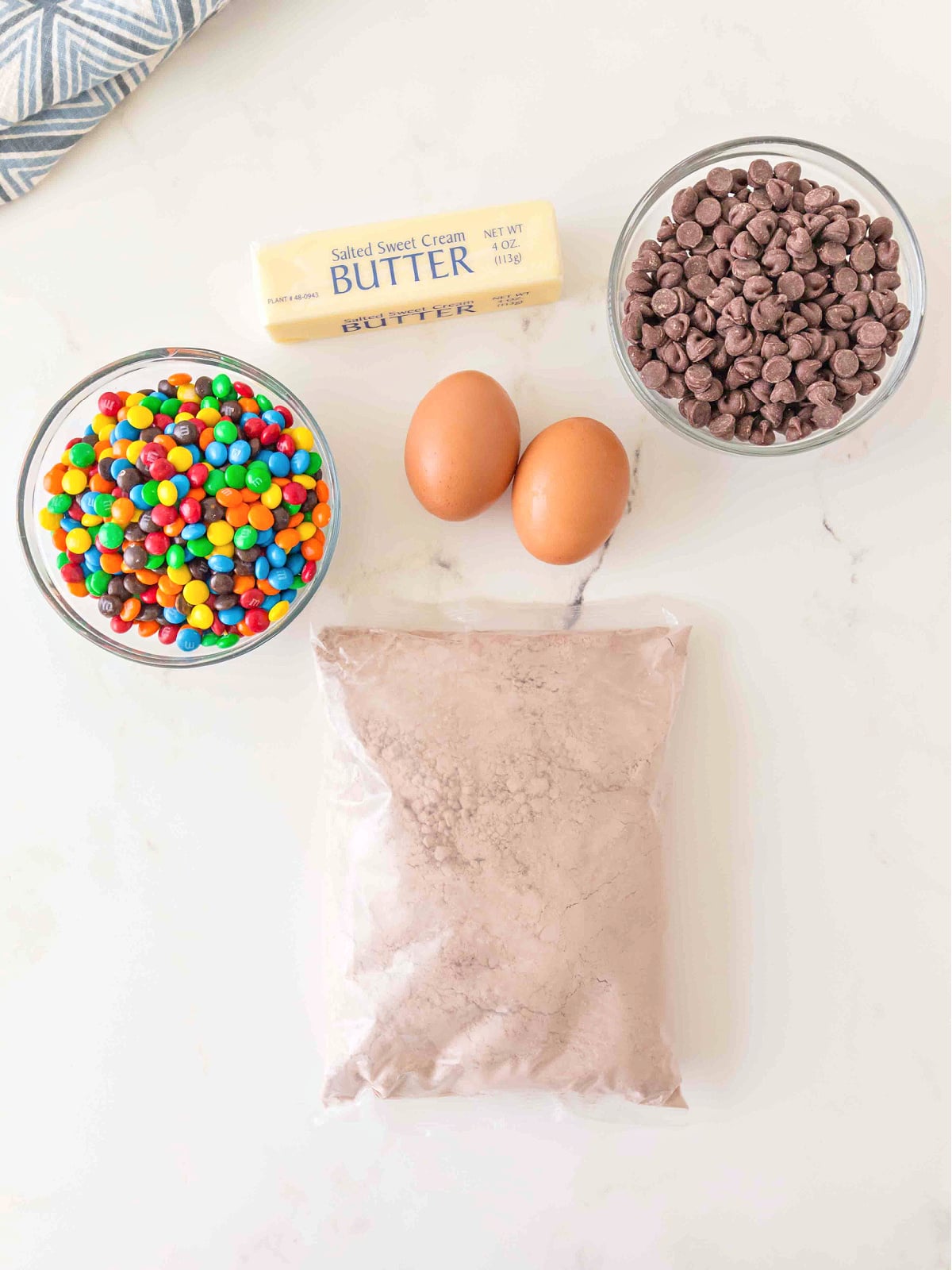 Brownie ingredients including cake mix, eggs, butter, chocolate chips, and M&M candies arranged on a countertop.