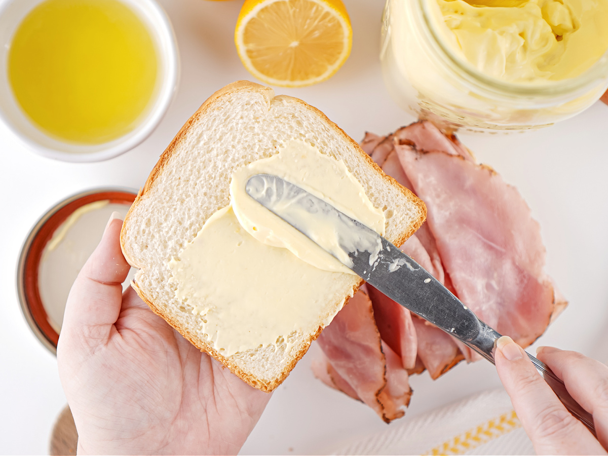 Homemade mayonnaise being spread onto white bread for a ham sandwich.