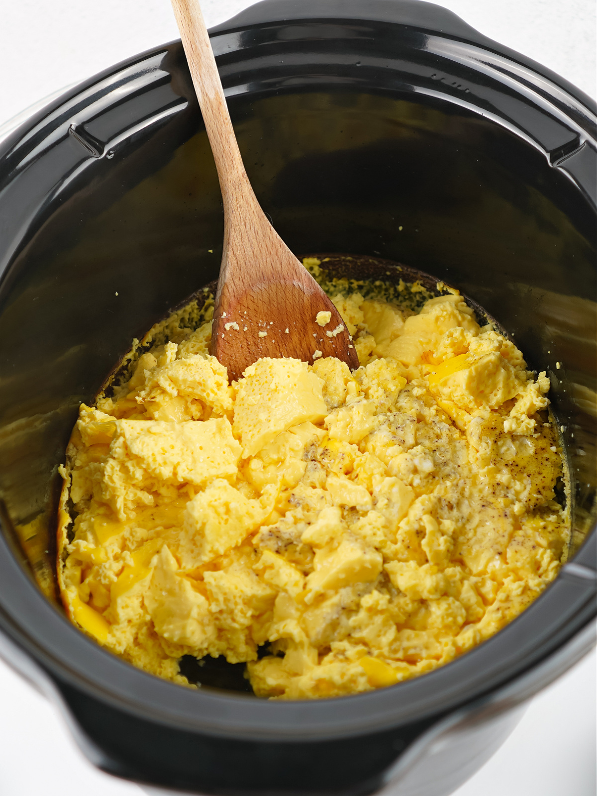 Wooden spoon lifting soft, fluffy scrambled eggs from a crockpot after cooking.