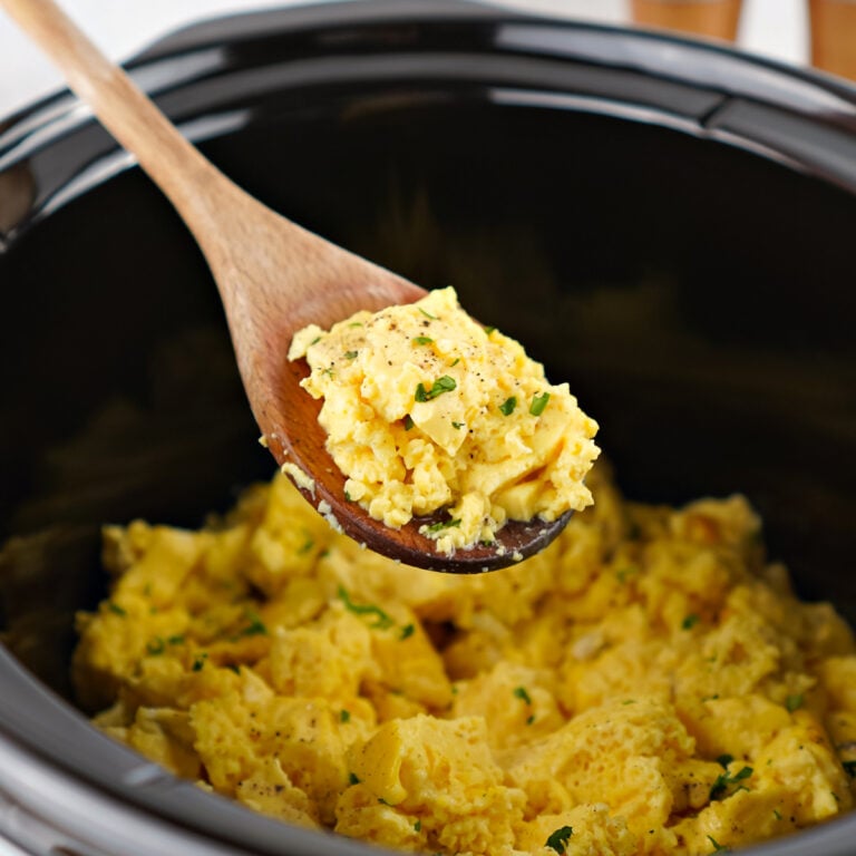 Wooden spoon stirring soft, fully cooked scrambled eggs inside a black crockpot.