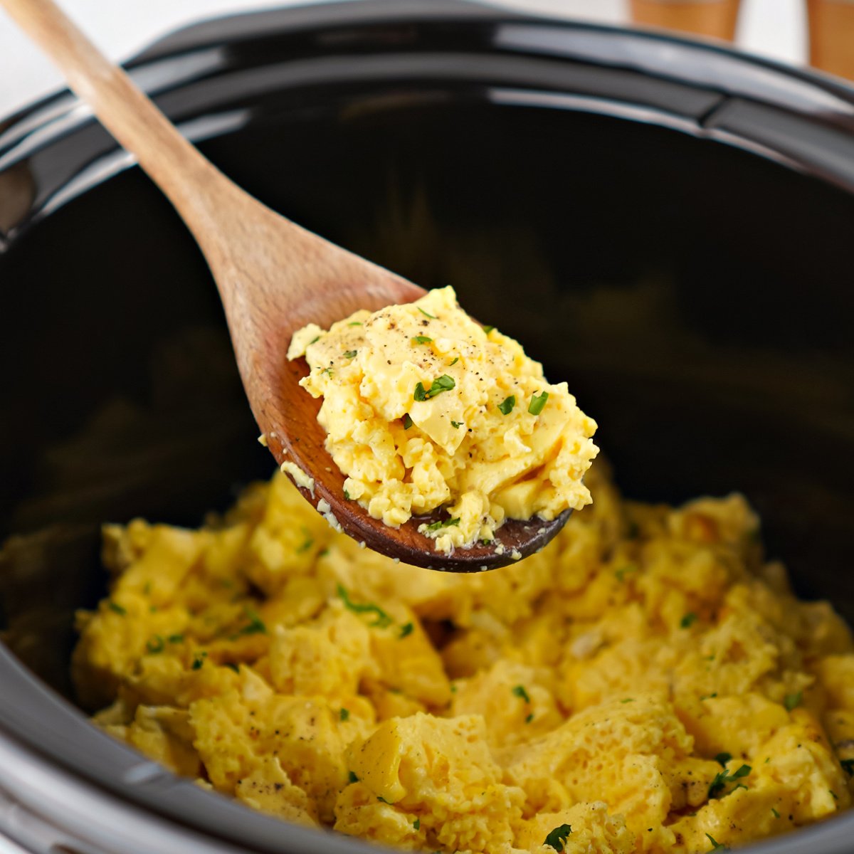 Wooden spoon stirring soft, fully cooked scrambled eggs inside a black crockpot.