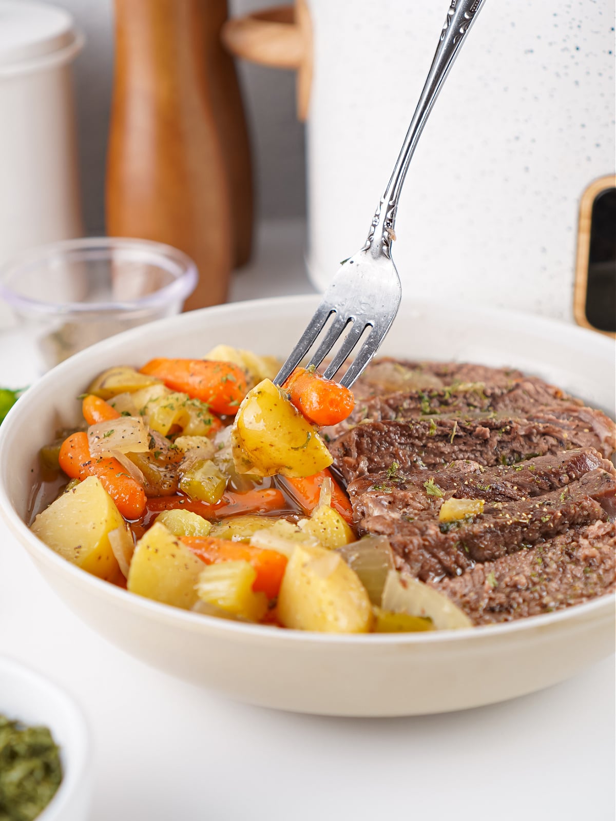 Crockpot seasoned pot roast served in a white bowl with carrots, potatoes, celery, and onions, with a fork lifting a bite of vegetables.