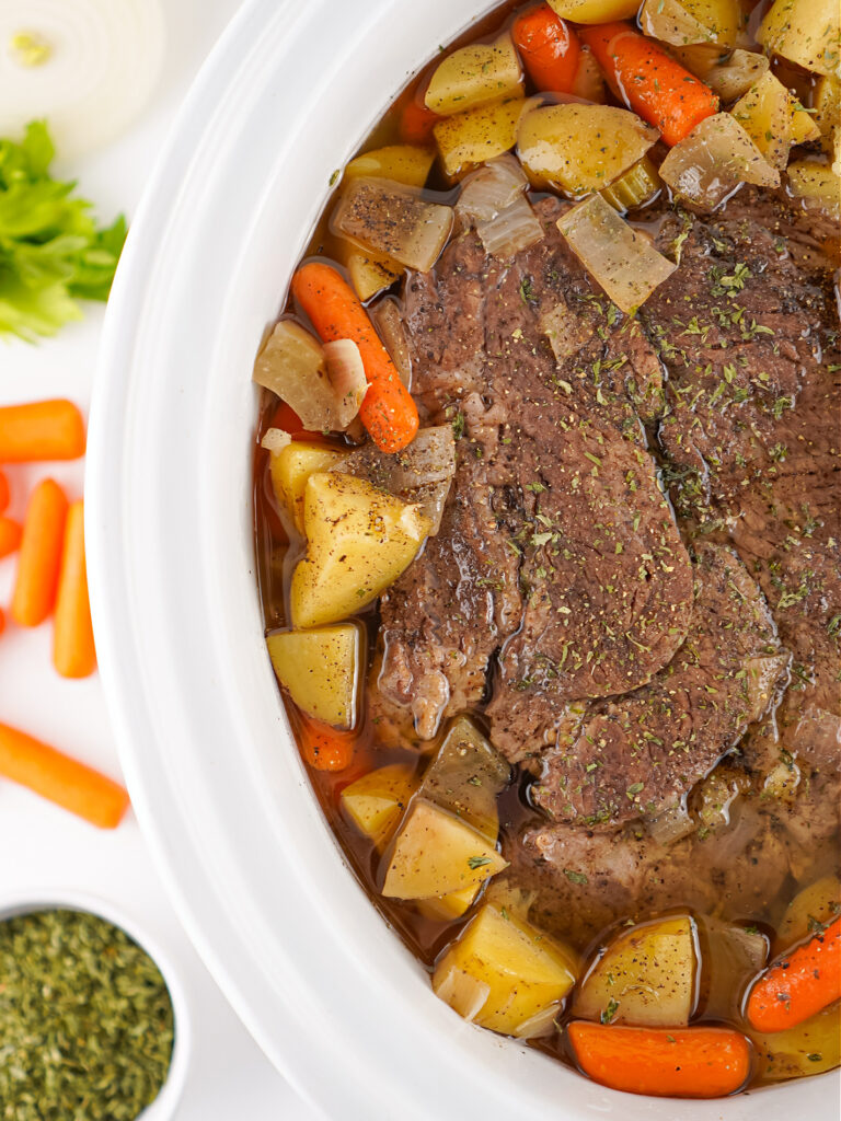 Top-down view of seasoned pot roast cooking in a slow cooker with carrots, potatoes, onions, and herbs in broth.