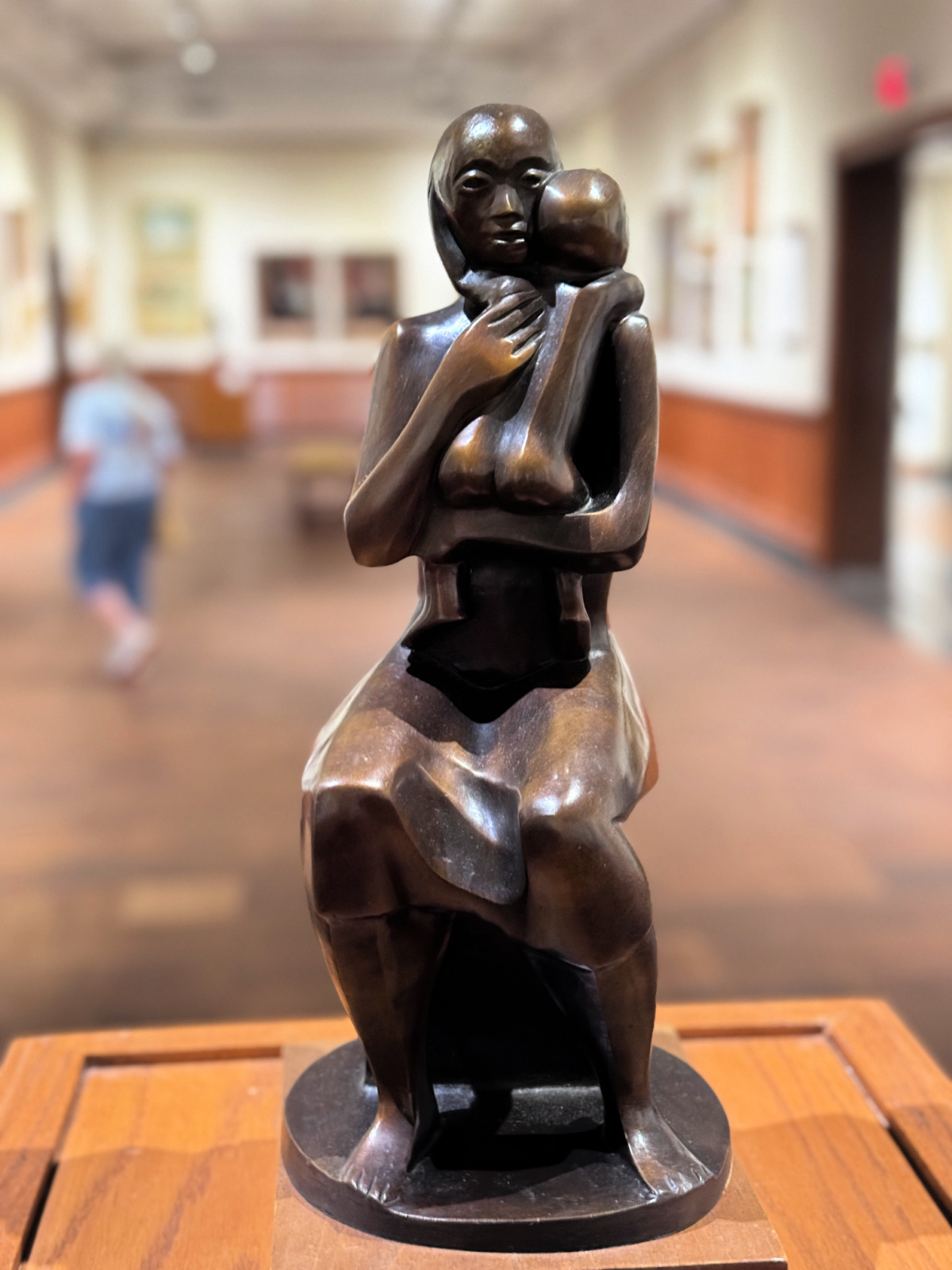 Bronze sculpture of a seated mother holding a child, displayed inside an art museum gallery with framed artwork in the background.