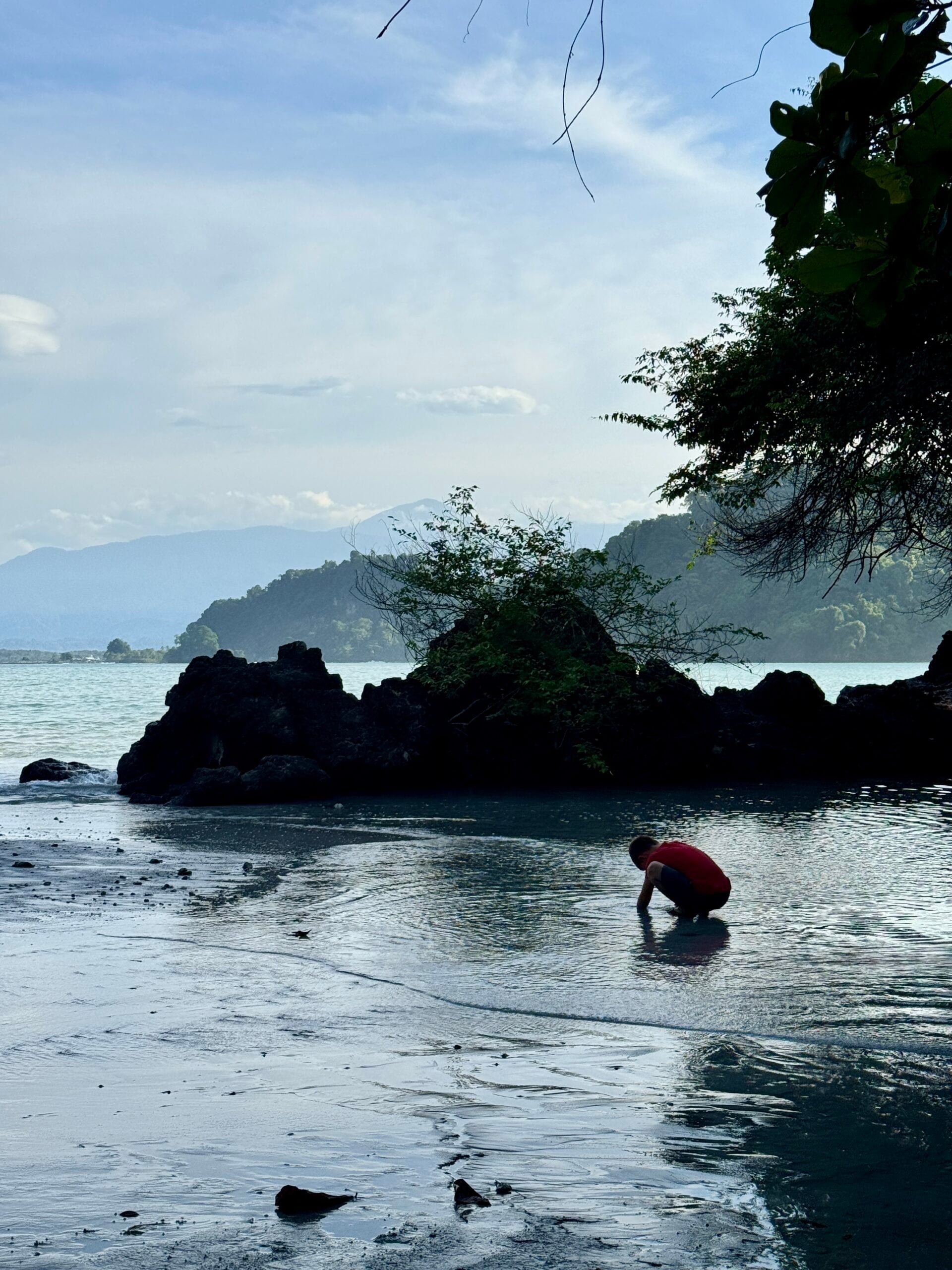 Child crouching in shallow water at Playa Biesanz in Manuel Antonio, Costa Rica, with rocky shoreline and jungle-covered hills in the background.